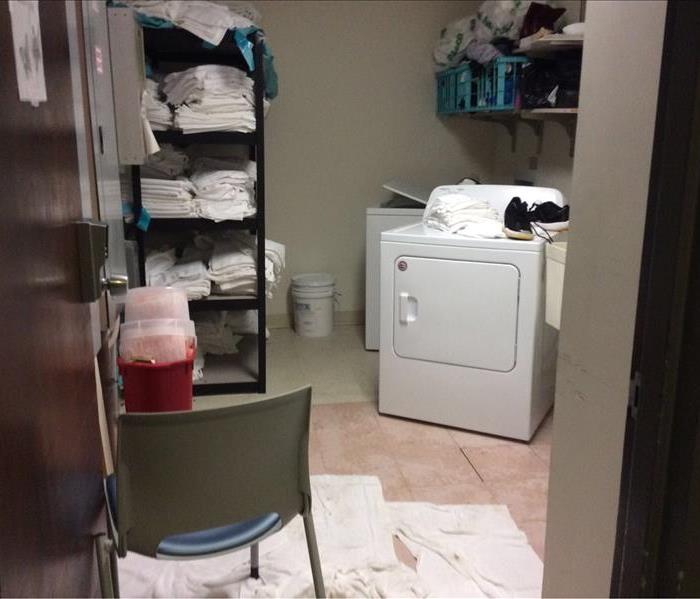 laundry room with towels on the floor and dryer pulled out from the wall. Towels on black shelving and electrical systems.